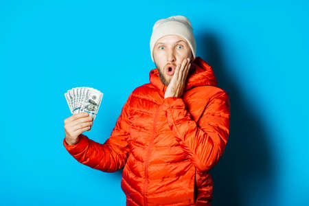 Man with a surprised face in a hat and a red jacket parka holds money in his hands on a blue background. Concept surprise, shock, win, lottery, sports betting, rich.の写真素材