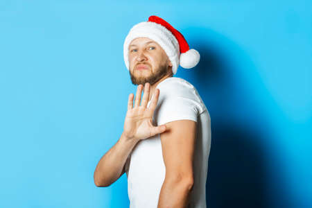 Man in a Santa Claus hat makes a repulsive gesture with his hands on a blue background. Concept no...