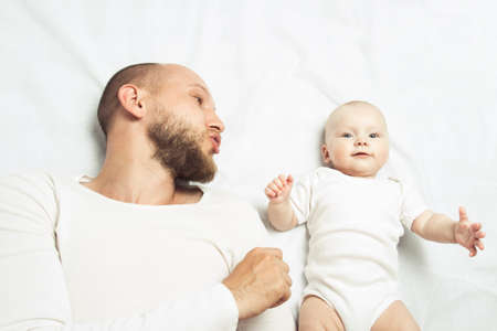 Young dad playing with little baby in white clothes on a bed with a white sheet. Flat lay, top view.