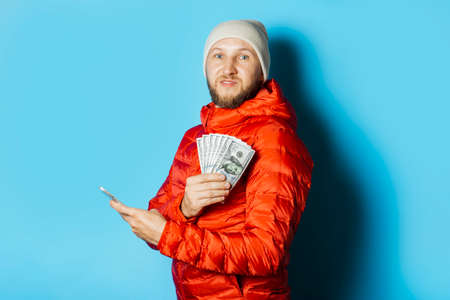 Man with a surprised face in a hat and a red jacket holds money and a mobile phone in his hands on a blue background. Concept surprise, shock, win, lottery, sports betting, betting, app.の写真素材