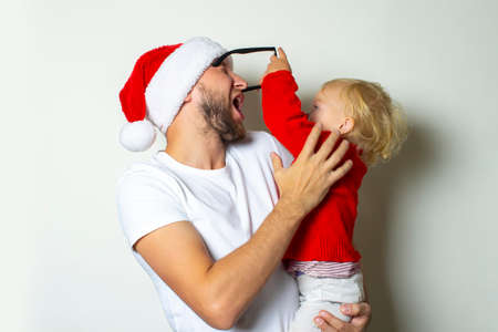 A young man in glasses and a Santa Claus hat plays with a child on a white background. Christmas concept, celebration with family, family holidays.の写真素材