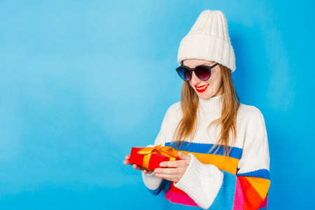 A young girl with a surprised face in glasses, a white hat and a sweater is holding a gift on a blue...