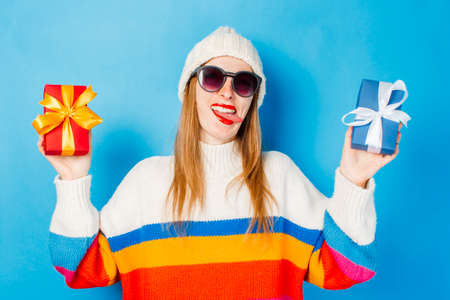 A young girl shows her tongue and grips in glasses, a white hat and a sweater holding a gift on a...
