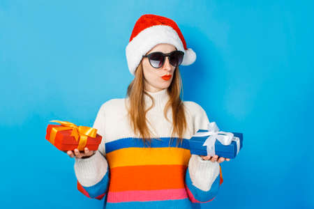 A young girl in a Santa Claus hat is holding gift boxes on a blue background. The concept of New Year and Christmas, gifts for the winter holidays, shopping at salesの写真素材