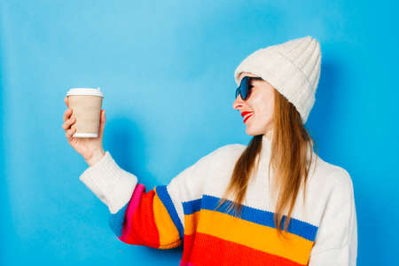 A young girl in glasses and a hat is holding a paper cup with coffee or tea and looking at it...