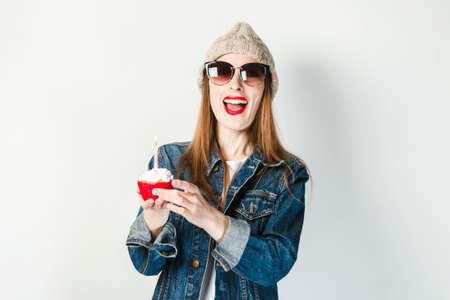 Young girl in a denim jacket, glasses and a hat holds a small cake with candles on a white...