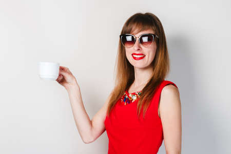 A young girl in a red dress holds in her hand a cup of coffee on a light background. Coffee shop...