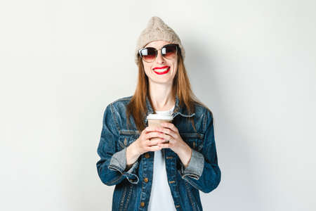 A young girl in a denim jacket, glasses and a hat is holding a paper cup of coffee or tea on a white...