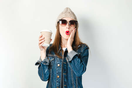 A young girl in a denim jacket, glasses and a hat is holding a paper cup of coffee or tea on a white background. Concept breakfast, coffee at work, coffee shop, rechargingの写真素材