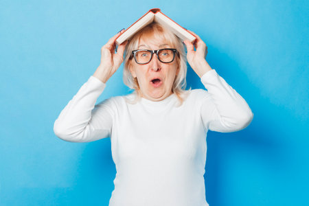 An old woman in glasses with a surprised face holding a book over her head against a blue background. Concept old lady reads books, education, book clubの写真素材
