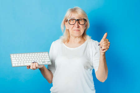 Old lady with glasses holds a keyboard on a blue background. Internet concept for old people, new technologies, computer learning by old people, technology.の写真素材