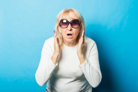 Old woman wearing sunglasses with a surprised face makes a gesture with her hands against a blue background. Concept of surprise, shock, unexpected news.の写真素材