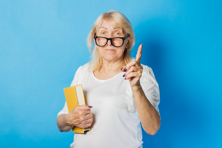 Old woman with glasses holds a book in her hands and shows a hand gesture on a blue background. Concept Knowledge and power of reading and education.の写真素材