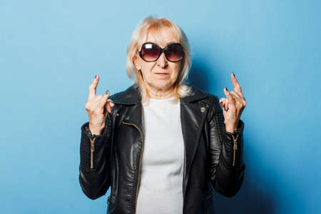 Old lady with glasses and a leather jacket is showing a gesture with her hands against a blue...