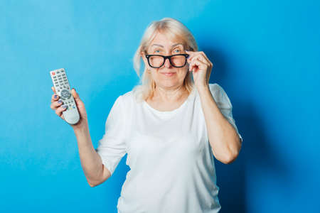 Old woman is holding a TV remote on a blue background. Concept television for the elderly, the study of new technologies, TV series and urgent keys, cable television, high quality.の写真素材