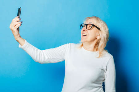 Old woman with glasses takes a selfie on a blue background. The concept of new technologies.