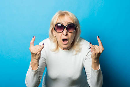 Old woman wearing glasses with a surprised face makes a gesture with her hands against a blue background. Concept rock, old rocker, goat gesture.の写真素材