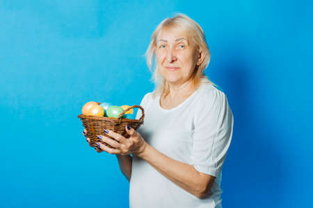 Old woman holds in her hands a basket of Easter eggs with me against a blue background. Easter celebration concept.の写真素材