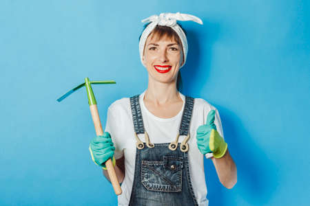 Young woman wearing gardener clothes is holding a tool against a blue background. Concept gardener, fertilizer and garden tools, high quality.の写真素材