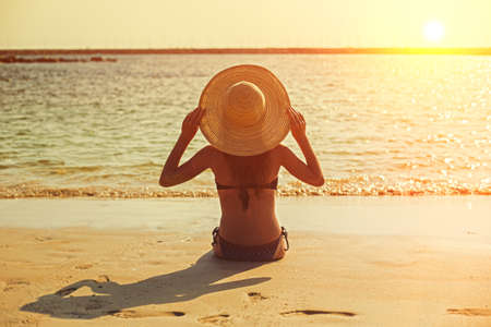 Young girl at sunset on the beach sits with her back to the sea in a hatの写真素材