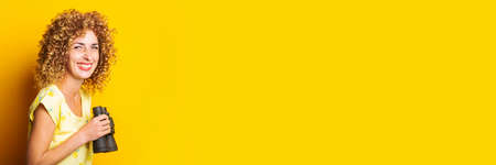 cheerful smiling curly girl holding binoculars in hands on a yellow background. Banner
