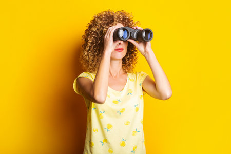 curly young woman looks through binoculars on a bright yellow backgroundの写真素材