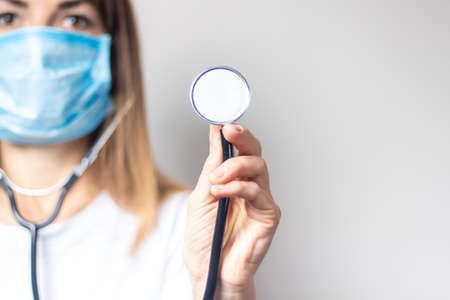 Young woman in a medical mask holds a stethoscope on a light background. Banner. Concept doctor,...