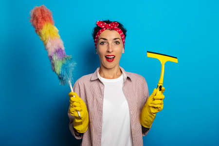 Surprised shocked young woman in gloves holding glass scraper and dust brush on blue backgroundの写真素材