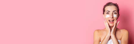 beautiful young woman applies cream to her face on a pink background. Banner