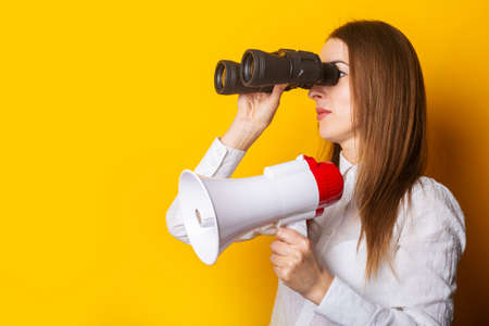Friendly young woman holds a megaphone in her hands and looks through binoculars on a yellow background. Hiring concept, help wanted. Banner.の写真素材
