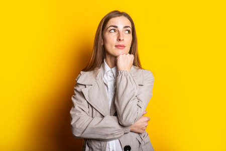 Young woman in a coat holds a hand under her chin and looks to the side against a yellow background. Banner.の写真素材