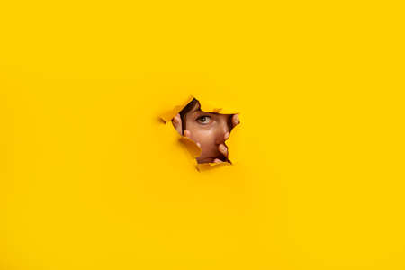 Young woman peeks out of a hole in the wall on a yellow background. Banner.