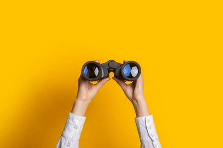female hands hold black binoculars on a bright yellow background.の写真素材