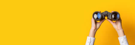 female hands hold black binoculars on a bright yellow background. Banner.の写真素材