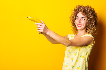 smiling curly young woman holding a banana pistol on a yellow background.の写真素材