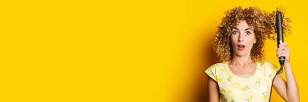 shocked girl with curly hair straightens her hair with a hair straightener on a yellow background....