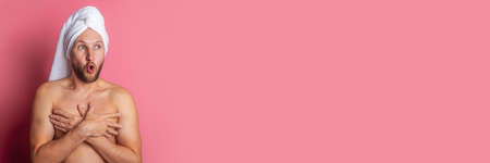 surprised naked young man with a towel on his head, covering his chest with his hands on a pink...