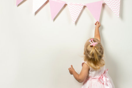 Little child hangs a festive garland of flags on a light background. Banner. Party, birthday, holiday, decoration concept.の写真素材