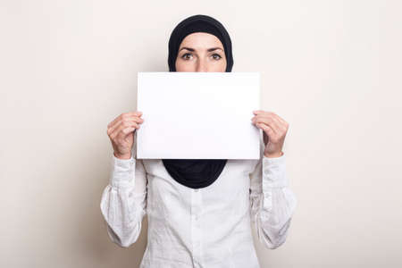 Young Muslim woman in a white shirt and hijab covers her face with a blank sheet of paper. Place for your text. Banner.の写真素材