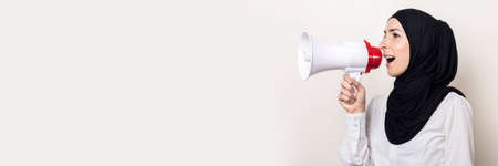Muslim young woman in hijab holds a megaphone in her hands and shouts into it on a light background....