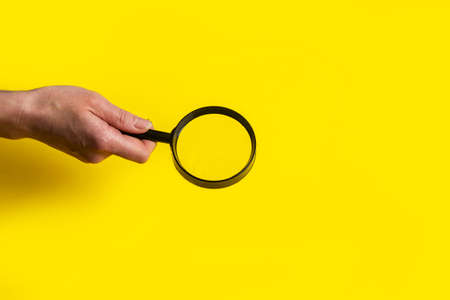female hand holds a magnifying glass loupe on a yellow background.