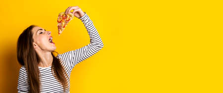 young woman eating a slice of hot fresh pizza on a yellow background. Banner.の写真素材