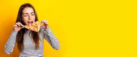 young woman bites a slice of hot fresh pizza on a yellow background. Banner.