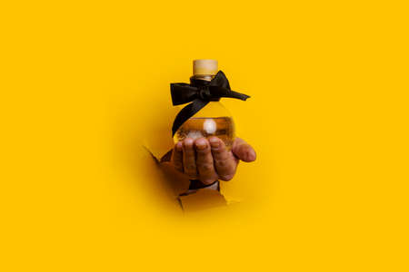 woman's hand holds a bottle with oil perfume in a torn hole on a yellow background.の写真素材