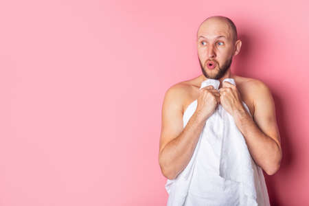 Surprised young man looks to the side, covering his bare chest with a towel against a pink background.の写真素材