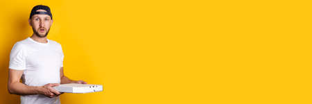 Surprised young man in a baseball cap holding a pizza box on a yellow background. Banner.の写真素材