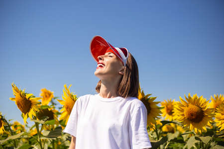 Young woman with closed eyes in red sun visor and white t-shirt looks at the sky on a sunflower field.の写真素材