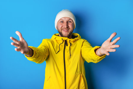 Joyful young man in a yellow jacket holds his palms in front of him meeting a friend on a blue...