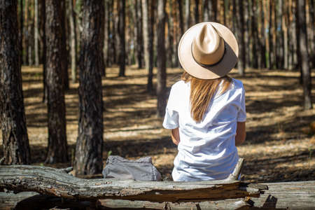 Young woman tourist in hat and t-shirt sits on a log during a halt in the forest.