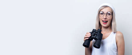 Cheerful surprised young woman holding binoculars looks to the side on a light background. Banner.の写真素材